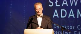 Na zdjęciu mężczyzna przemawiający z mównicy, ubrany w elegancki strój, w tle niebieskie tło z napisami.
