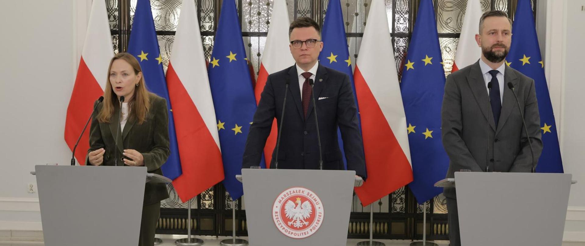 Minister Katarzyna Pełczyńska-Nałęcz wraz z marszałkiem Sejmu Szymonem Hołownią i wicepremierem, ministrem obrony narodowej Władysławem Kosiniakiem-Kamysz stoją za mównicami, za ich plecami flagi Polski i UE