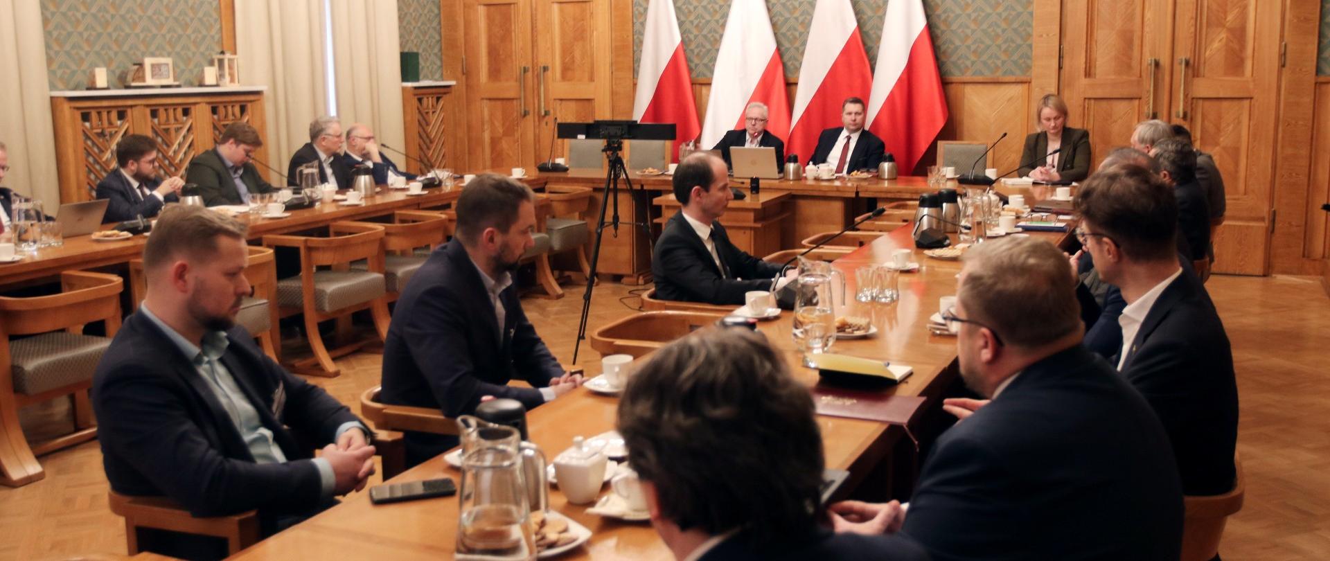W dużej sali z wyłożonymi drewnem ścianami, przy ustawionych w czworokąt drewnianych stołach siedzi kilkanaście osób, u szczytu stołu minister Czarnek i mężczyzna w garniturze, za nimi polskie flagi.