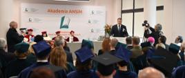 Widok z tyłu na salę, za mównicą stoi minister Czarnek i mówi do mikrofonu, za nim za białym stołem siedzą cztery osoby w czerwonych ceremonialnych strojach, przed nim wypełniona ludźmi sala.