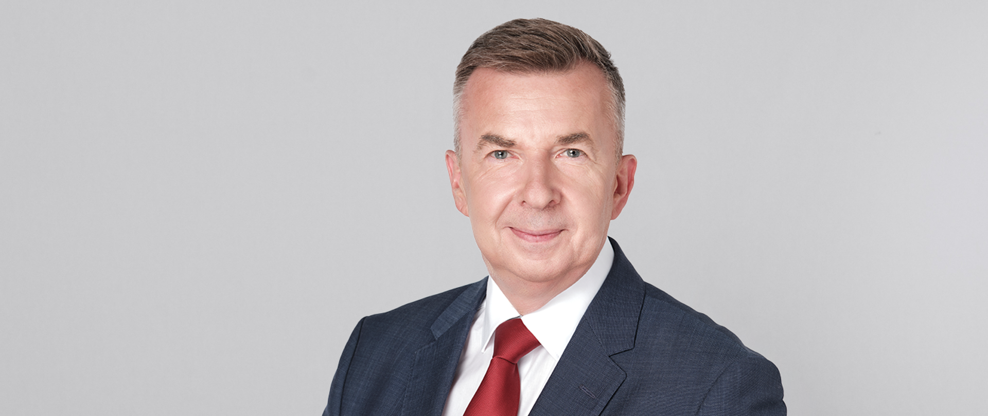 Minister Dariusz Wieczorek w szarym garniturze i czerwonym krawacie, zdjęcie na jasnoszarym tle.
