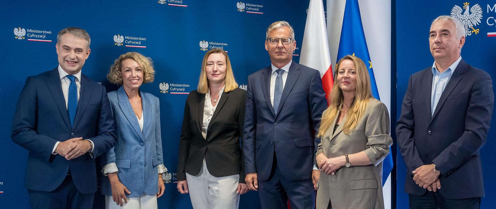 Na zdjęciu widzimy osoby, biorące udział w Konferencja prasowa - dotyczącej płatności w mObywatelu. Od prawej strony na zdjęciu znajduje się wicepremier i minister przedstawiciel miasta Bydgoszczy - Pan Janusz Popielewski, następna osoba to pani Ewa Szczecińska-Bedryj, Zekretarz Miasta Złotoryja, pan Mirosław Czekaj, prezes BGK, Joanna Erdamn, prezeska Fundacji Polska Bezgotówkowa, Karolina Matysiak z Centralnego Ośrodka Informatyki oraz wicepremier Krzysztof Gawkowski 