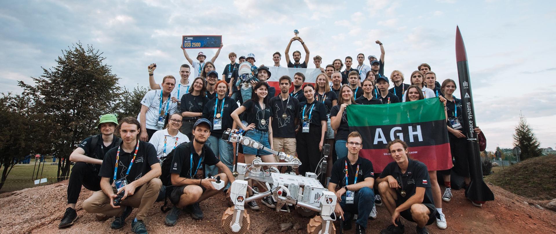 Pochmurny dzień, na piaszczystym pagórku duża grupa ludzi, trzymają zielono-czarno-czerwoną flagę z napisem AGH, przed nimi stoi czterokołowy robot.