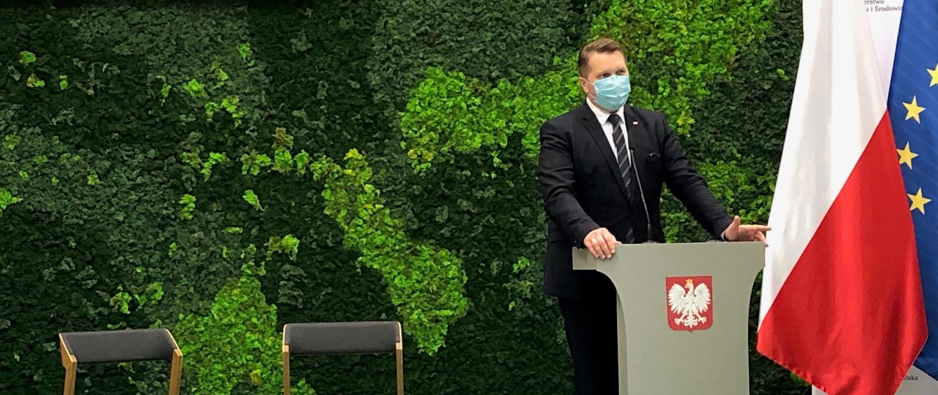 Minister Edukacji i Nauki przemawia stojąc za mównicą z godłem Polski, w tle zielona ściana imitująca rośliny oraz flagi Polski i Unii