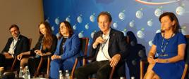 Wiceminister Bernacki siedzi obok kilku osób i mówi do mikrofonu, za nimi niebieska ściana.
