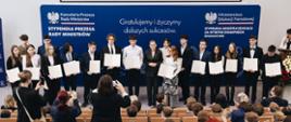 Druga grupa stypendystów pozuje na scenie z Barbarą Nowacką na tle niebieskiej ścianki informacyjnej. Minister stoi w środku, uśmiechając się i trzymając splecione dłonie. Przed sceną widać siedzącą publiczność oraz osoby robiące zdjęcia telefonami.