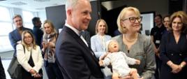 W sali z ciemnoszarymi ścianami stoi grupa ludzi, z przodu mężczyzna w czarnym garniturze i kobieta w szarej sukience i okularach trzymają manekin małego dziecka, za nimi szpitalne łóżko z żółto-różową pościelą.