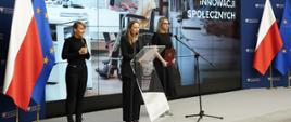 Minister funduszy i polityki regionalnej Katarzyna Pełczyńska-Nałęcz i wiceministra Monika Sikora podczas konferencji prasowej poświęconej inkubatorom na innowacje społeczne, panie minister stoją na scenie, obok nich stoi tłumaczka języka migowego