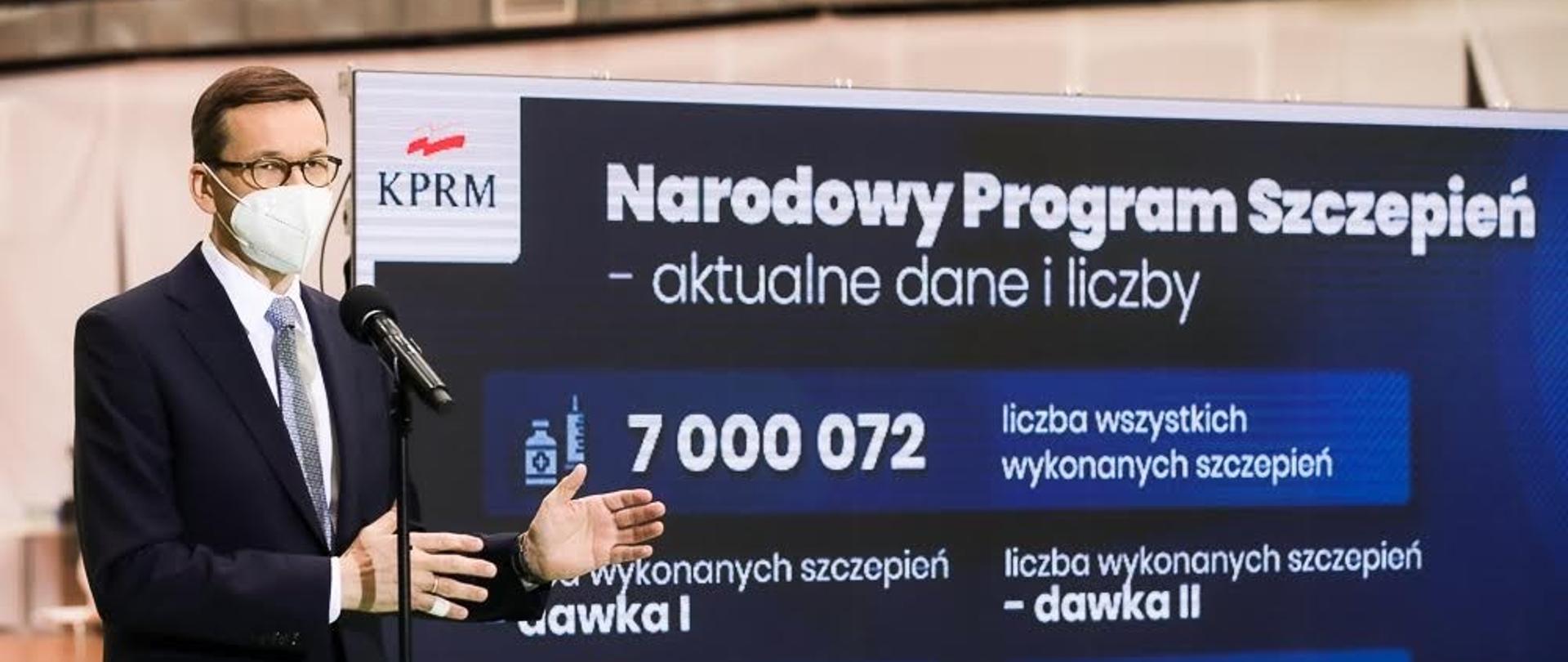 Premier Mateusz Morawiecki wskazuje na ekran z napisem Narodowy Program Szczepień - aktualne dane i liczby.