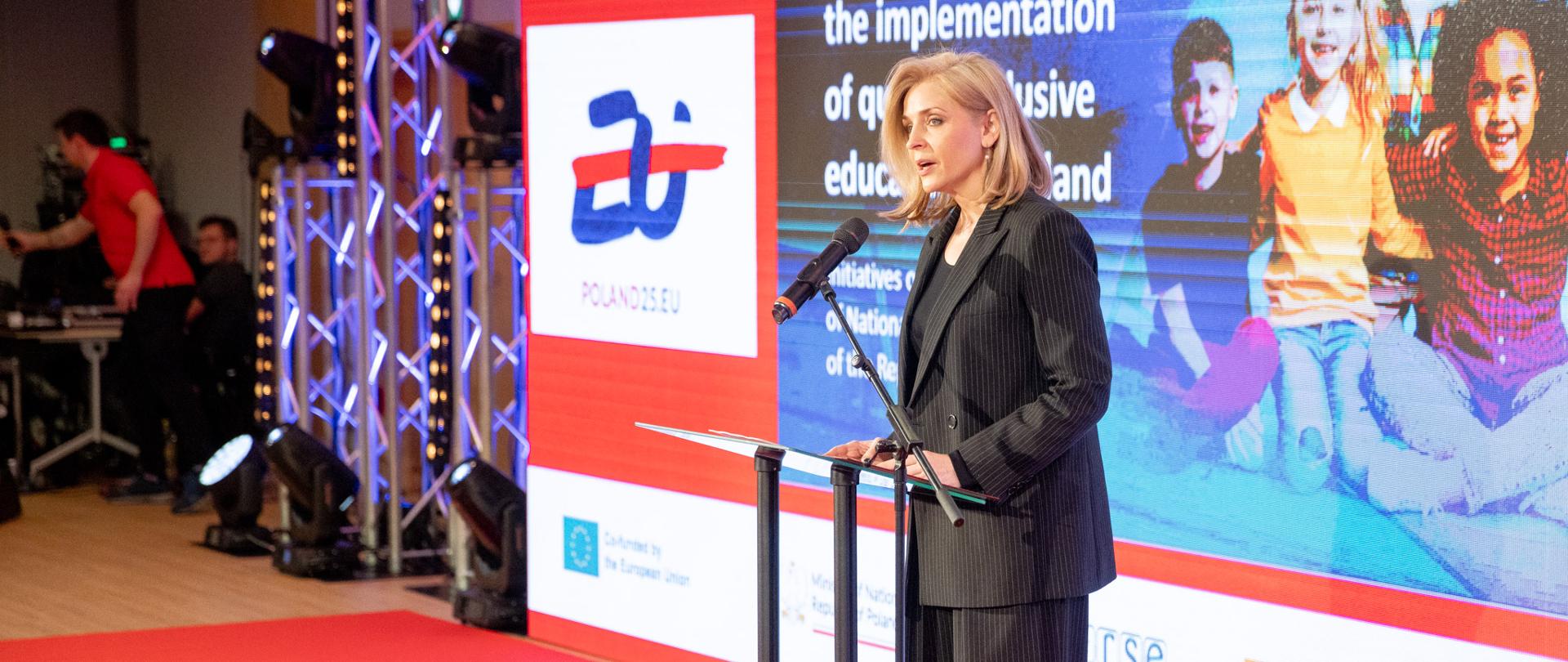 Let’s Make Inclusion Happen – inauguracja konferencji na temat edukacji włączającej z udziałem wiceminister Joanny Muchy, wiceminister w czasie wydarzenia przemawia przy mównicy do mikrofonu, ubrana w czarny garnitur w paski