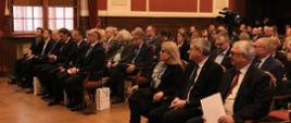Zdjęcie z boku na salę, na ustawionych w rzędy krzesłach siedzą elegancko ubrani ludzie, obok krzeseł stoją papierowe torby, w tle czerwono-beżowa ściana i okno.