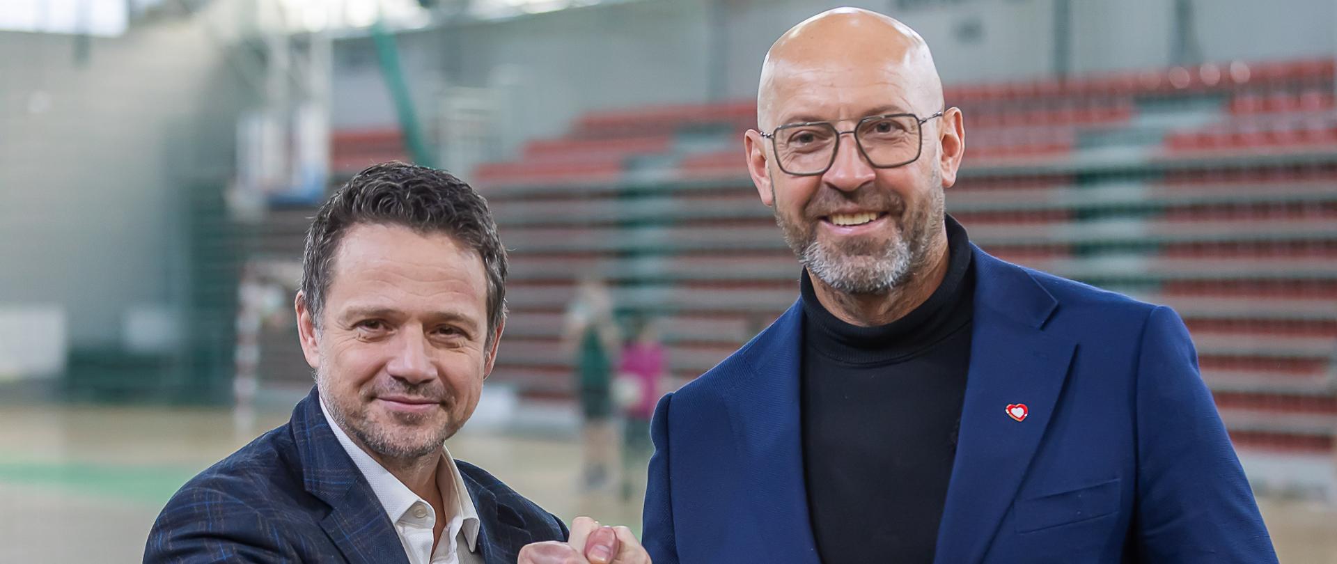 Zdjęcie prezentujące uścisk dłoni między prezydentem Warszawy Rafałem Trzaskowskim i ministrem sportu i turystyki Jakubem Rutnickim. Zdjęcie zrobione na hali sportowej.