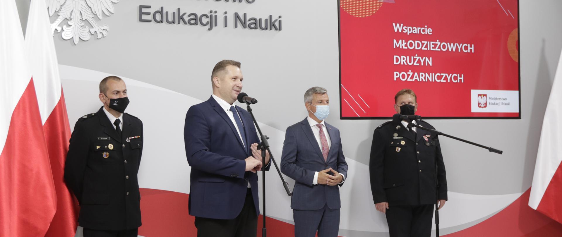 Minister Przemysław Czarnek stoi przy mikrofonie i przemawia. Po jego lewej stronie strażak w mundurze, po prawej mężczyzna w garniturze oraz strażak w mundurze. Panowie mają na twarzach maseczki. Po prawej stronie ekran, na którym widnieje napis Wsparcie Młodzieżowych Drużyn Pożarniczych. W tle biało-czerwone flagi oraz godło i napis Ministerstwo Edukacji i Nauki. 
