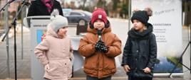 Troje małych dzieci stoi na dworze, za nimi mównica przy której stoi ubrana na czarno kobieta, dalej składany daszek z napisem Zakrzówek.