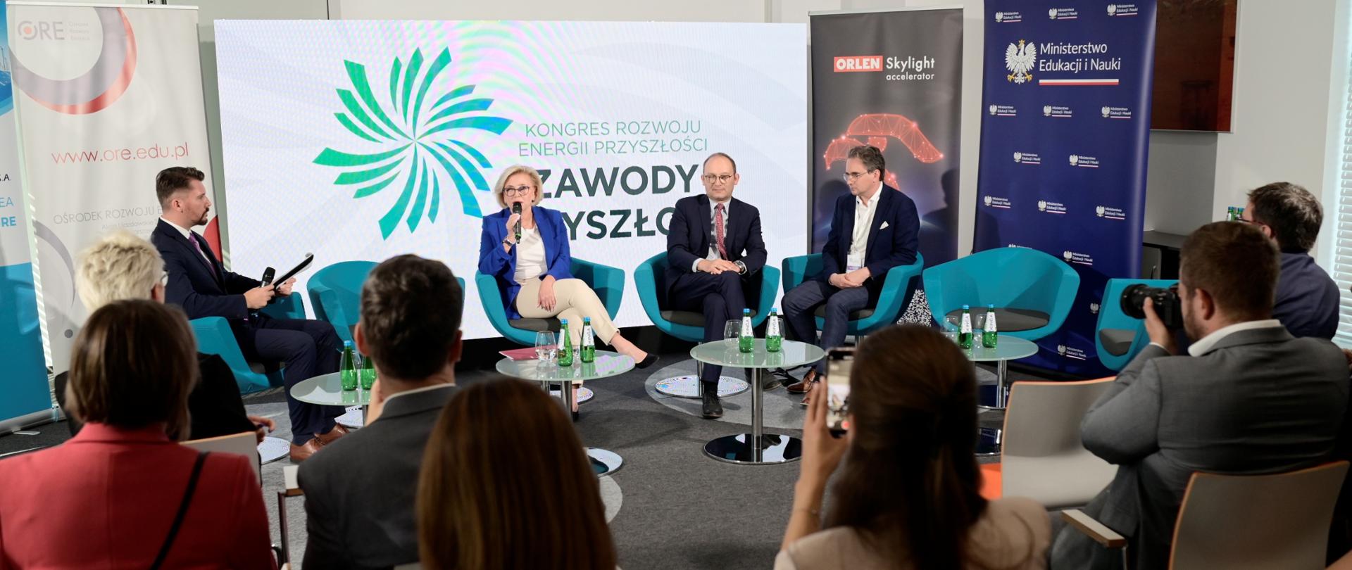 Widok z tyłu na salę, wiceminister Machałek w niebieskiej marynarce siedzi na niebieskim fotelu i mówi do mikrofonu, na fotelach po obu stronach siedzi trzech mężczyzn w garniturach, za nimi ścianka z napisem Zawody przyszłości, przed nimi na krzesłach siedzą ludzie.