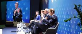 Wiceminister funduszy i polityki regionalnej Jan Szyszko siedzi w fotelu, po jego prawej stronie siedzą pozostali uczestnicy panelu, w głębszej części planu telebim na którym wyświetlony jest wiceminister J. Szyszko 