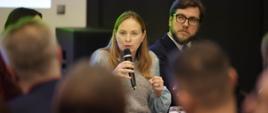 Minister funduszy i polityki regionalnej Katarzyna Pełczyńska-Nałęcz siedzi przy stole i przemawia do mikrofonu trzymanego w dłoniach.