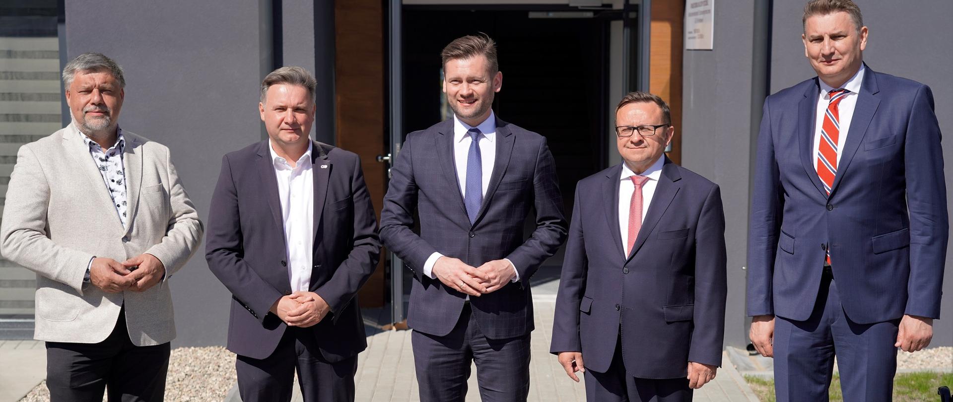 Minister Kamil Bortniczuk stoi przed wejściem do budynku wraz z gośćmi biorącymi udział w uroczystości otwarcia internatu sportowego w Centralnym Ośrodku Sportu Cetniewo