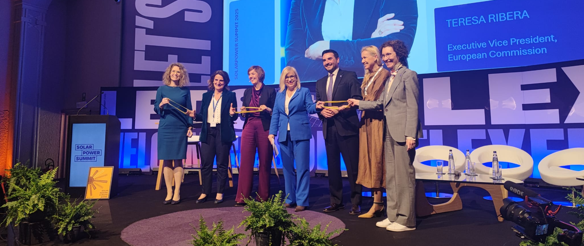 Ministra Paulina Hennig-Kloska w trakcie otwarcia konferencji z okazji 40-lecia działalności SolarPower Europe, organizacji zrzeszającej podmioty z branży fotowoltaicznej w Europie. Ministra stoi na scenie w towarzystwie przedstawicieli branży. Uśmiechnięci, podają sobie dokumenty i pozują do zdjęcia. 