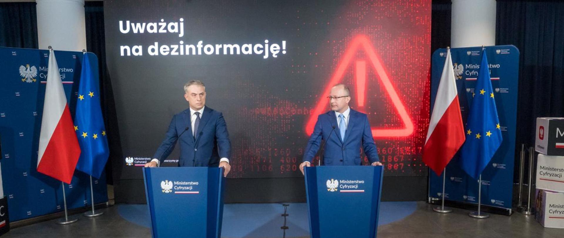 Dwie osoby stoją przy mównicy podczas konferencji prasowej. W tle widać napis "uważaj na dezinformację".