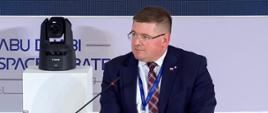 Wiceminister siedzi na białym fotelu, przed nim mikrofon na długim stojaku, za nim jasnoszara ściana.
