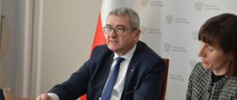 Wiceminister Murdzek siedzi za biurkiem i parezy w ekran stojącego przed nim laptopa, obok niego ubrana w marynarkę w szarą kratkę kobieta pisze na kartce, za nimi polska flaga.