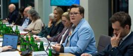Zbliżenie na kobietę (Katarzyna Lubnauer) w jasnoniebieskiej marynarce i okularach, siedzącą przy stole z dokumentami i mikrofonem, prawdopodobnie wygłaszającą przemówienie lub prezentację podczas posiedzenia. W tle widoczny jest ekran z napisem "KOMPAS JUTRA".