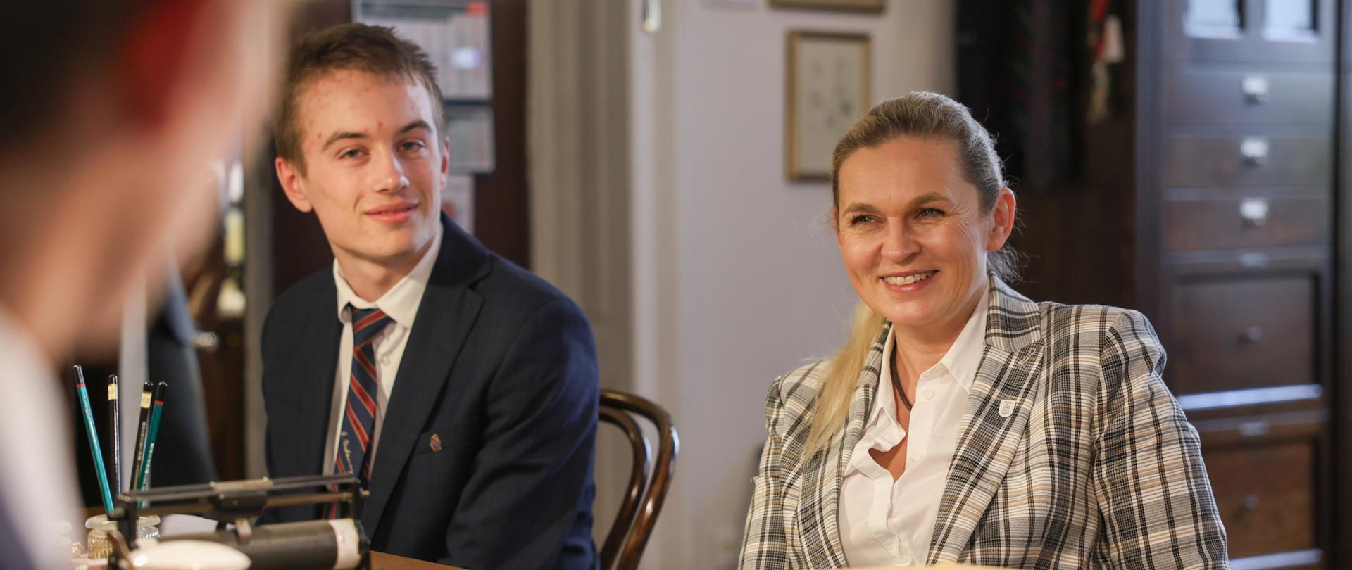 Minister Edukacji siedzi przy stole i rozmawia z przedstawicielami samorządu uczniowskiego. Obok niej siedzi uczeń. 