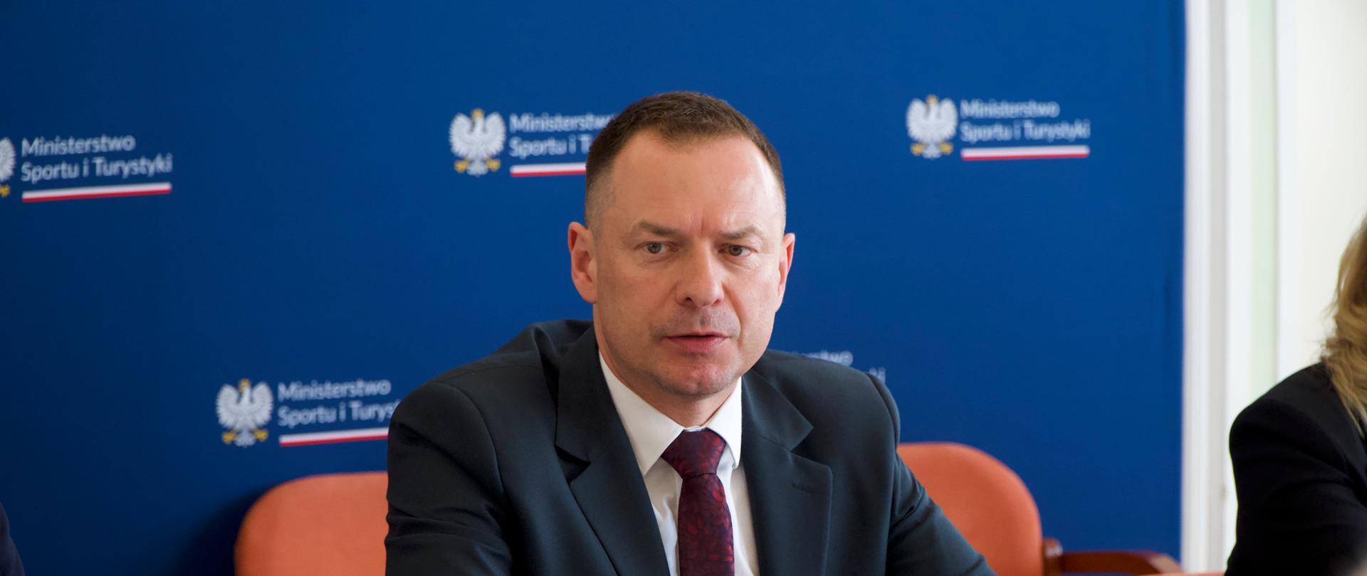 Posiedzenie zespołu ds. szlaków turystycznych. Na zdjęciu Piotr Borys siedzi na tle niebieskiej ścianki z logiem ministerstwa