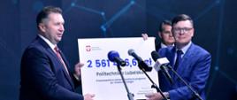 Minister Czarnek i drugi mężczyzna stoją przed mikrofonami i trzymają w rękach duży symboliczny czek z kwotą której nie widać - zasłaniają ją mikrofony.