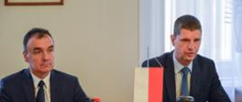 Za drewnianym stołem siedzi wiceminister Piontkowski i mężczyzna w garniturze, przed nimi mała polska flaga, za nimi żółta ściana i okno.