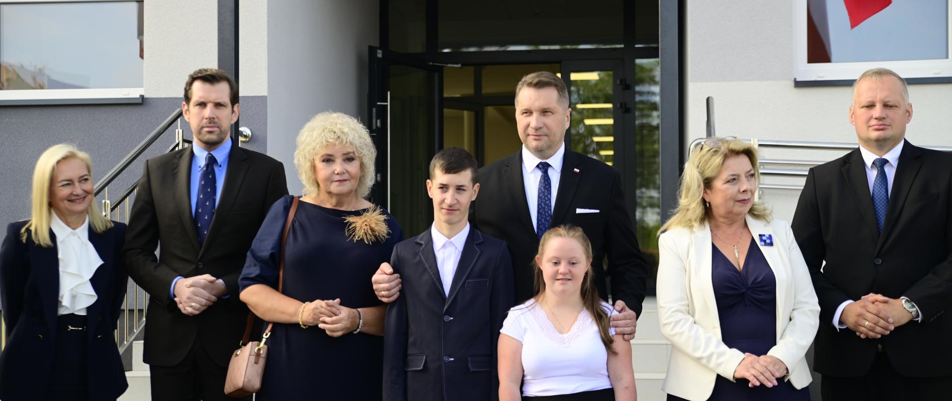 Przed wejściem do budynku stoi minister Czarnek otoczony przez kilka osób.