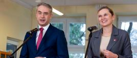 Portretowe ujęcie Krzysztofa Gawkowskiego i Barbary Nowackiej stojących przy mikrofonach. Oboje są uśmiechnięci i patrzą przed siebie. W rozmytym tle widać fragment sali lekcyjnej i uczniów.