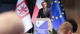 Wiceminister Jacek Karnowski stoi w środku na sali i mówi do mikrofonu.