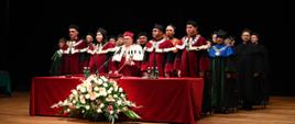 W zaciemnionej sali przy przykrytym czerwonym materiałem stole stoją ludzie w ceremonialnych czerwonych strojach z białymi futrzanymi kołnierzami.