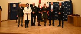 Grupa ludzi, czterech mężczyzn i trzy kobiety, stoi na drewnianej podłodze, za nimi na żółtej ścianie duży czerwony krzyż i ścianka z napisami KUL.
