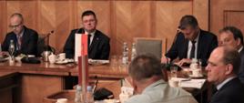 Sześciu mężczyzn siedzi przy stole, jeden pracuje na laptopie, drugi pisze na kartce, trzech słucha, jeden mówi. Na stole filiżanki, woda, ciastka i owoce