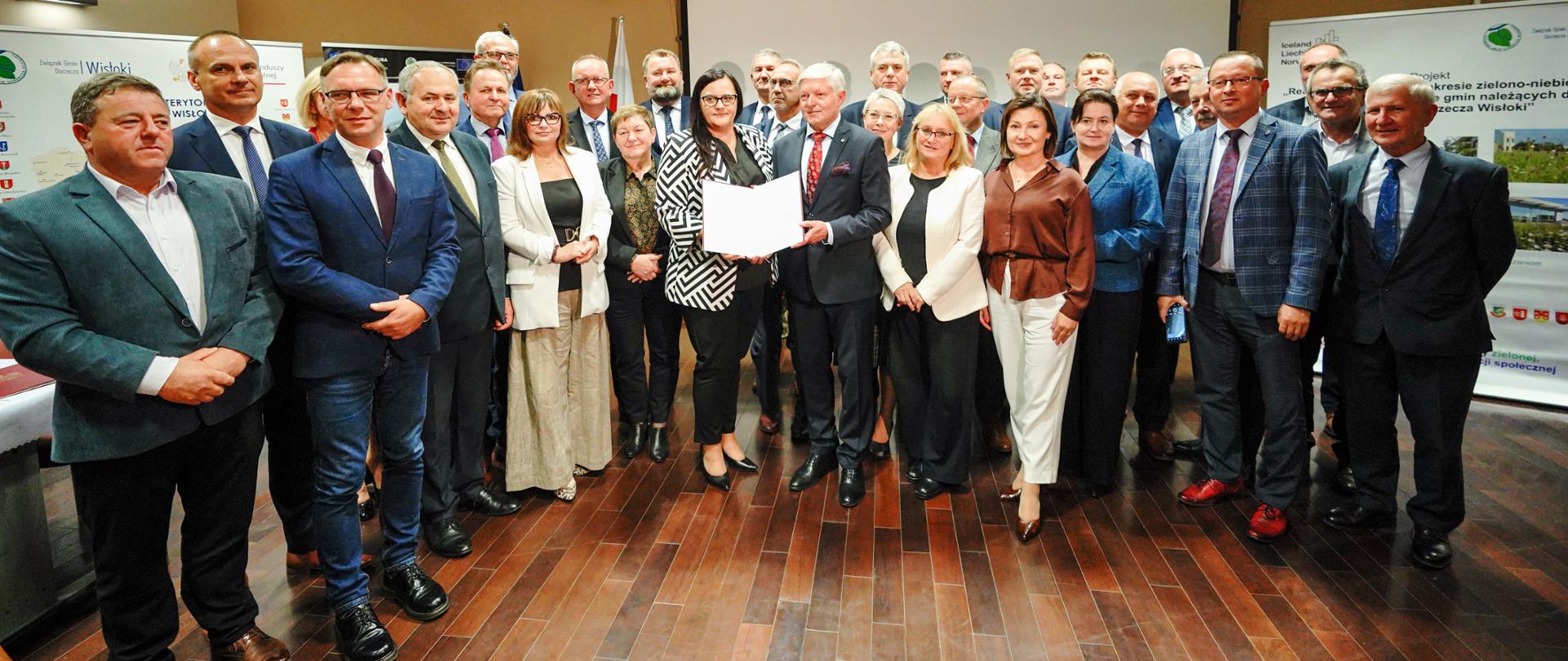 W sali grupa osób pozuje do zdjęcia. Dwójka z nich prezentuje otwarty dokument.
