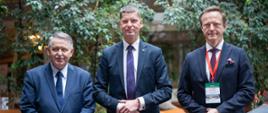 Wiceminister Piontkowski stoi obok dwóch mężczyzn w garniturach, za nimi brązowe fotele i duże zielone rośliny w doniczkach.