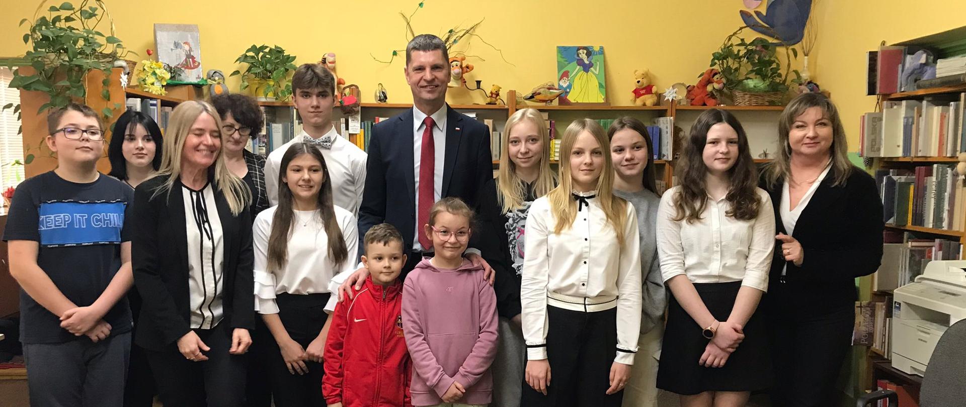 Pod żółtą ścianą stoi grupa młodzieży i dzieci, w środku grupy wiceminister Piontkowski, za nimi półki z książkami.