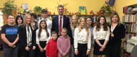 Pod żółtą ścianą stoi grupa młodzieży i dzieci, w środku grupy wiceminister Piontkowski, za nimi półki z książkami.