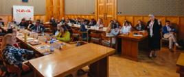 W sali ze ścianami wyłożonymi drewnianymi panelami przy ustawionych w czworokąt stołach siedzi dużo ludzi, w tle pod ścianą baner z napisem NAWA.
