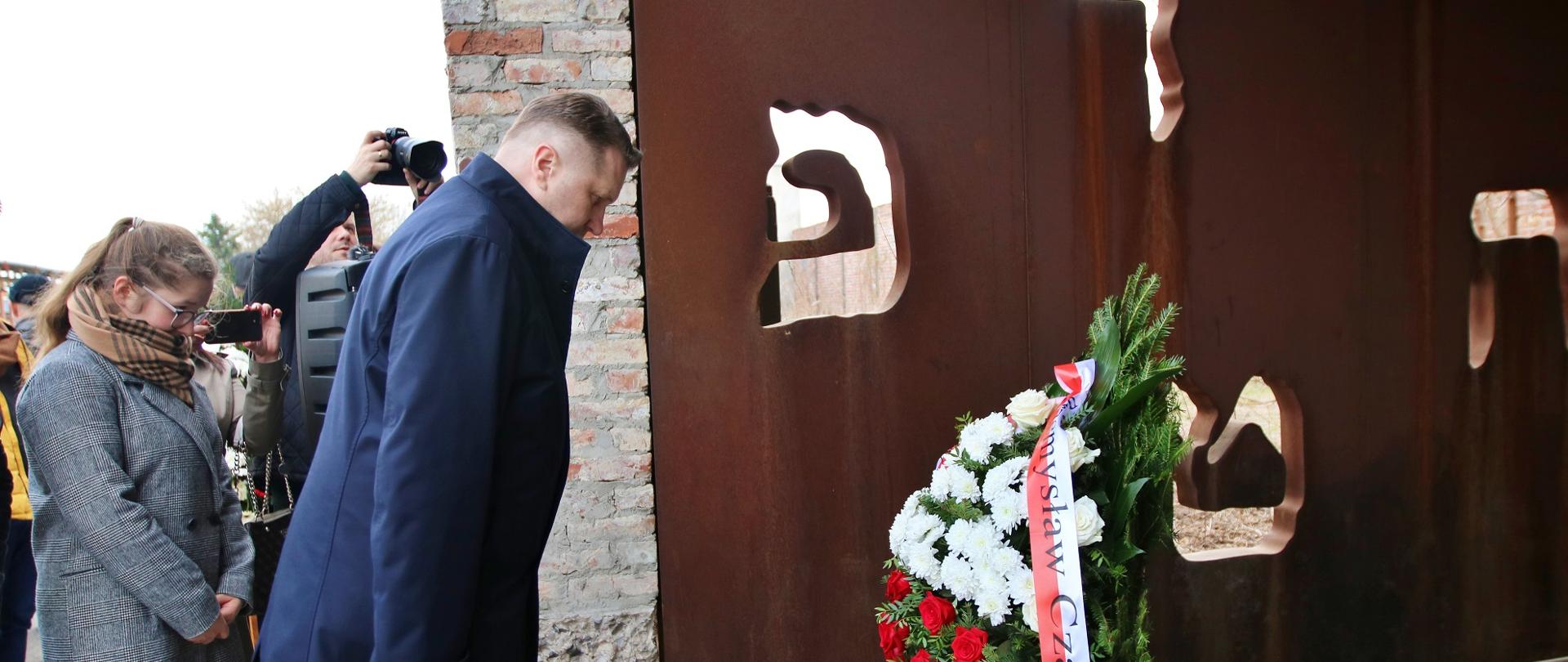 Minister Czarnek w czarnej kurtce pochyla się przed stojącym na podstawce biało-czerwonym wieńcem, za nim ceglana ściana, obok kobieta w szarym płaszczu.