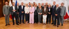 Pamiątkowe zdjęcie grupowe uczestników konferencji stojących w rzędzie na tle ścianki z logo oraz flag Polski i Unii Europejskiej. W centrum stoi Barbara Nowacka w różowym garniturze.