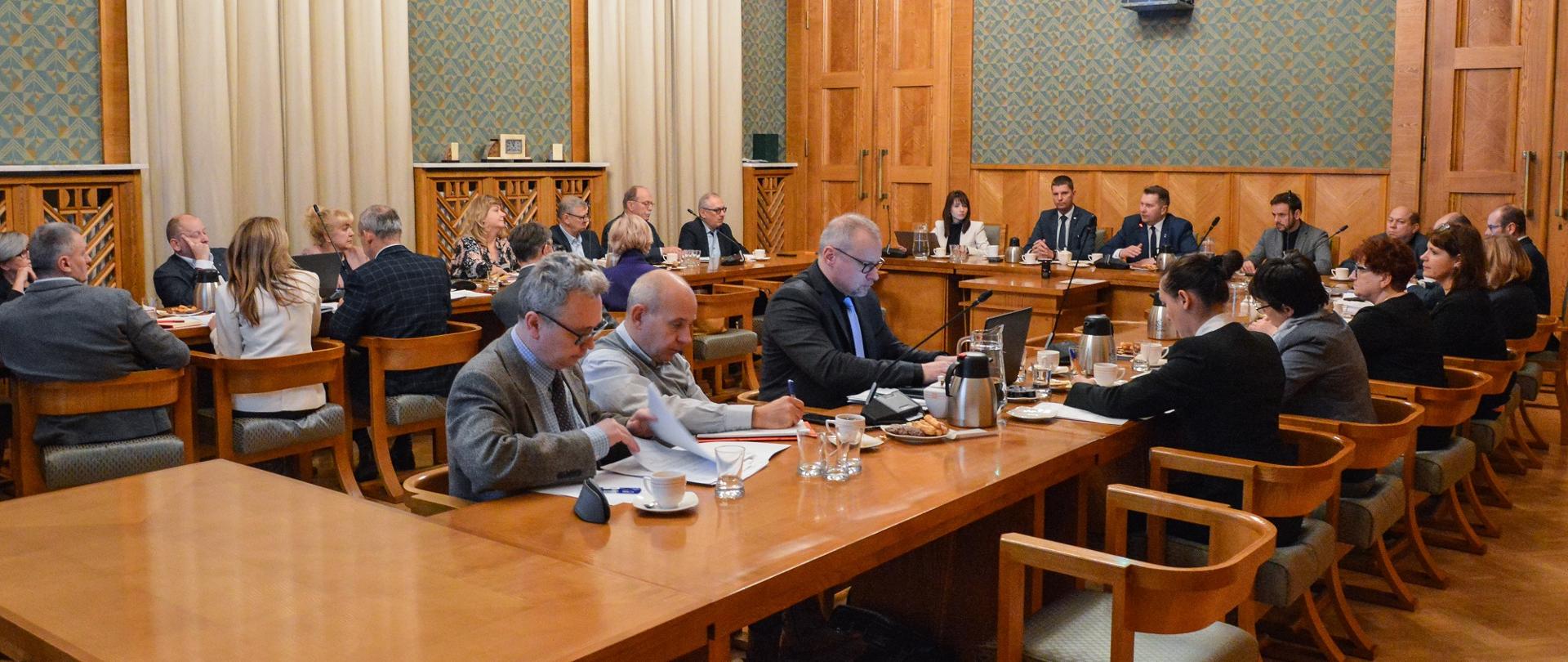 Duża sala ze ścianami w zielony wzorek, przy drewnianych stołach ustawionych w czworokąt siedzi dużo elegancko ubranych osób.