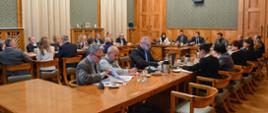 Duża sala ze ścianami w zielony wzorek, przy drewnianych stołach ustawionych w czworokąt siedzi dużo elegancko ubranych osób.