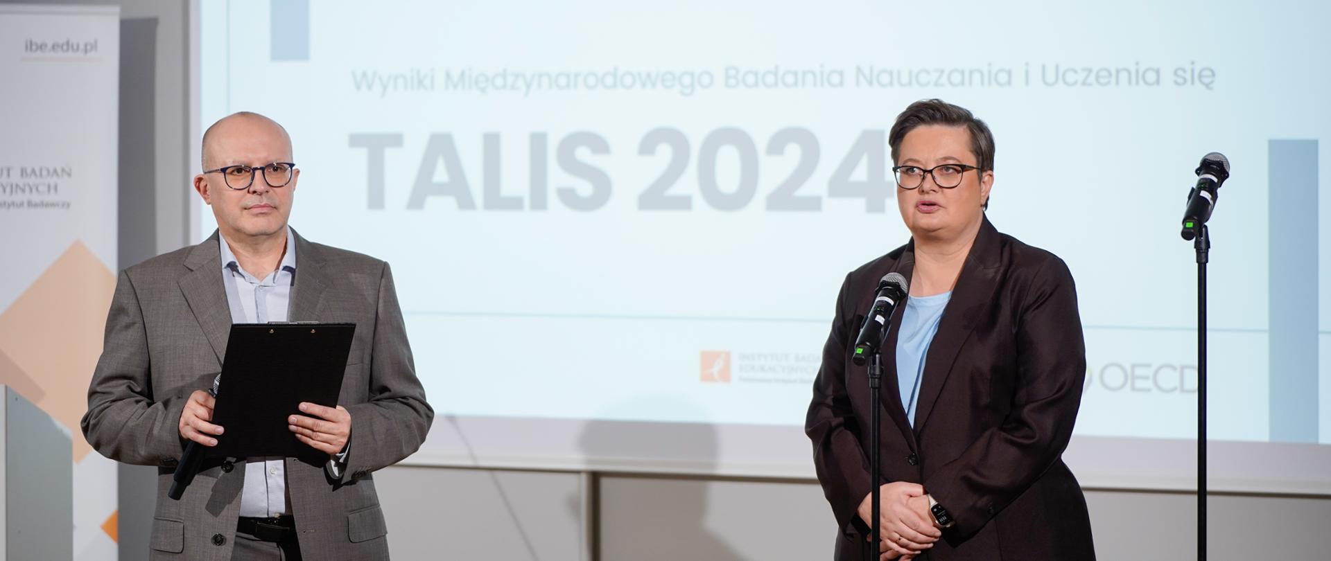 Dwóch prelegentów na scenie podczas konferencji dotyczącej wyników badania TALIS 2024. Mężczyzna w szarym garniturze trzyma czarną teczkę po lewej, a kobieta w ciemnym garniturze i okularach stoi przy mikrofonie po prawej. W tle widoczny jest ekran z tytułem: Nauczycielki i nauczyciele o pracy w zawodzie, Wyniki Międzynarodowego Badania Nauczania i Uczenia się oraz duży napis TALIS 2024."