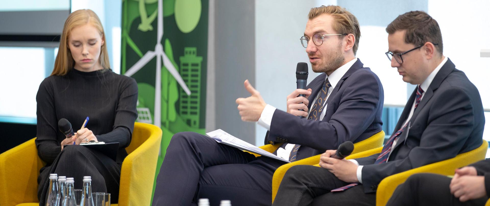 Grupa osób siedzi na fotelach i rozmawia. Przed nimi publiczność. Od lewej wiceminister Jan Szyszko z mikrofonem.