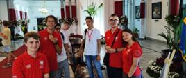 Grupa młodych ludzi w czerwonych i białych koszulkach stoi w dużym pomieszczeniu z kolumnami i obrazami na ścianach.