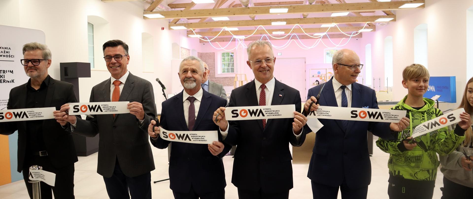 W dużej sali stoi w rzędzie grupa osób, pięciu mężczyzn w garniturach i dwoje dzieci w kolorowych bluzach, wszyscy trzymają kawałki wstęgi z napisami SOWA.